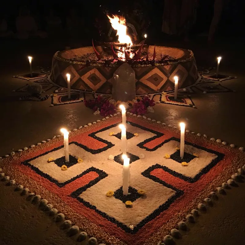 A ceremonial fire burning in a round, diamond-patterned hearth, with a quartz crystal, flowers and candles before it and a geometric floor design of seeds and grains