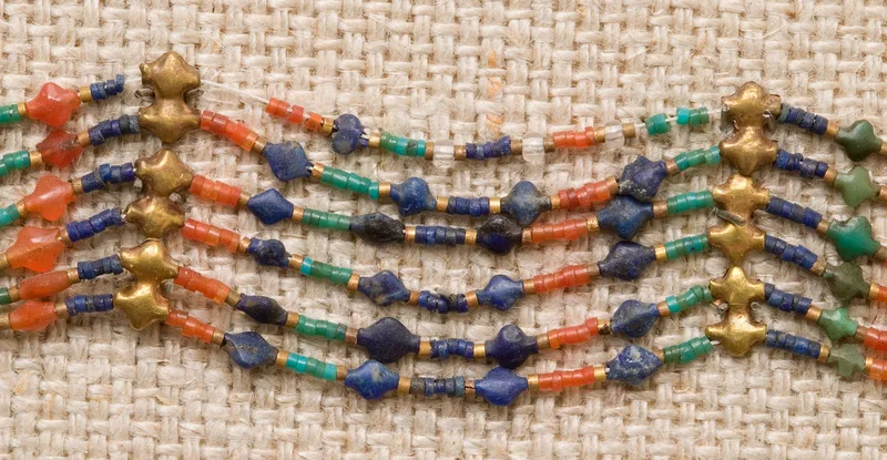 Detail of a bead girdle on linen: rows of four-lobed seed-shaped beads in gold, blue, red and green stone, strung between small cylinder beads