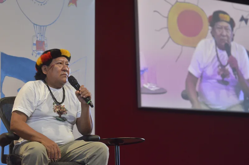 Davi Kopenawa, wearing a headdress of feathers, speaks into a microphone on a stage, with his own image projected on a screen behind him.