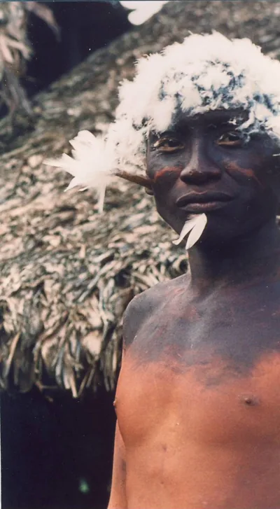 A young Waiká man wearing white down in his hair and dark body paint, standing before a thatched house