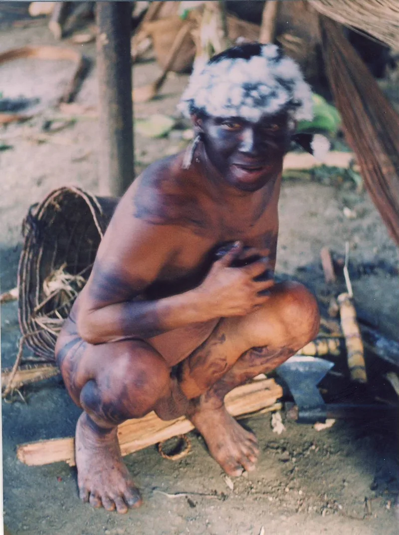 A Waiká man crouching, white down in his hair, his face and body painted dark