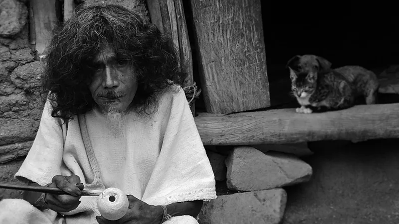 Black-and-white photograph of a long-haired man in a white tunic seated at a stone wall, working the stick in his poporo, with a cat behind him
