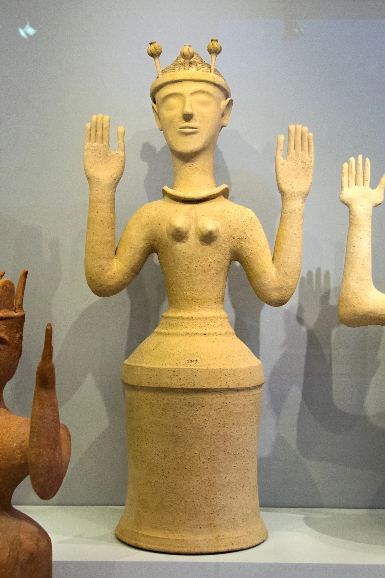 A pale terracotta female figure with both hands raised, a cylindrical skirt, and three pins topped with poppy capsules rising from her crown