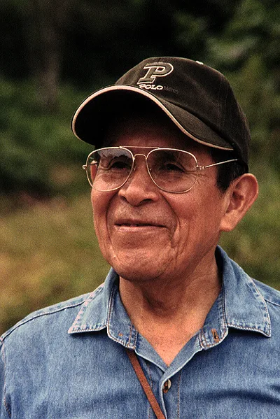 Pablo Amaringo in a denim shirt, glasses and a cap, smiling, outdoors among trees, in 2002.