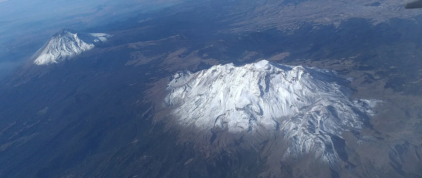 Iztaccíhuatl and Popocatépetl from the air. The weather workers Benítez Corona records are concerned with rain, with agriculture, and with waters held inside these mountains — a subterranean realm their dreams furnish with pipes, reservoirs and bells.