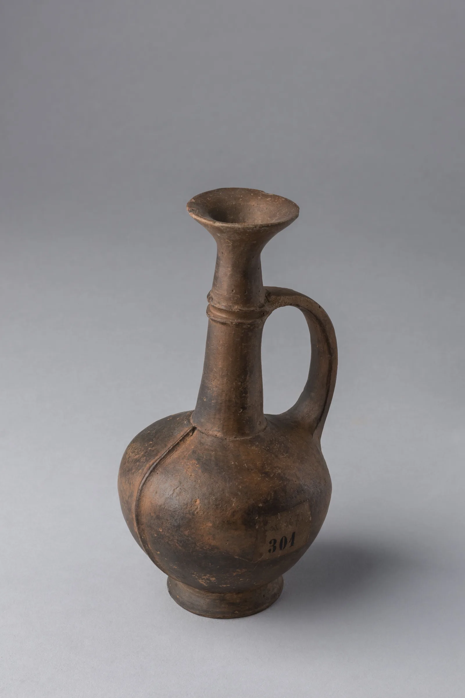 A brown clay juglet with a long narrow neck, a loop handle, a rounded body with raised lines and a ring base