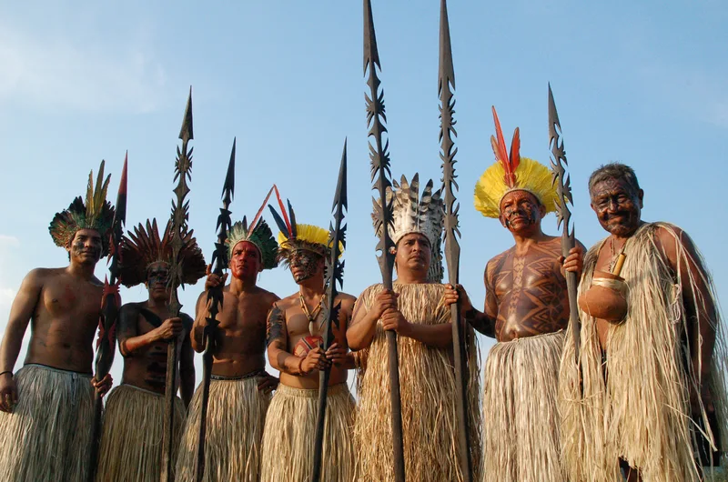 Eight Yawanawa men standing against a blue sky in feather headdresses and straw skirts, several with painted bodies, holding tall carved spears.
