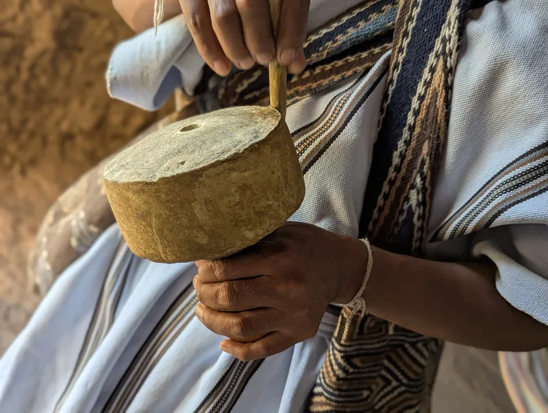 A gourd poporo held in a man’s hand, its mouth built up into a thick, pale crust of lime, with the wooden stick in it; he wears a white tunic and a woven mochila